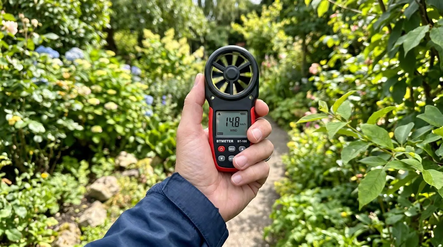 Gros plan sur un anémomètre portatif tenu à la main affichant une mesure de vitesse de vent, avec un jardin lumineux en arrière-plan flouté